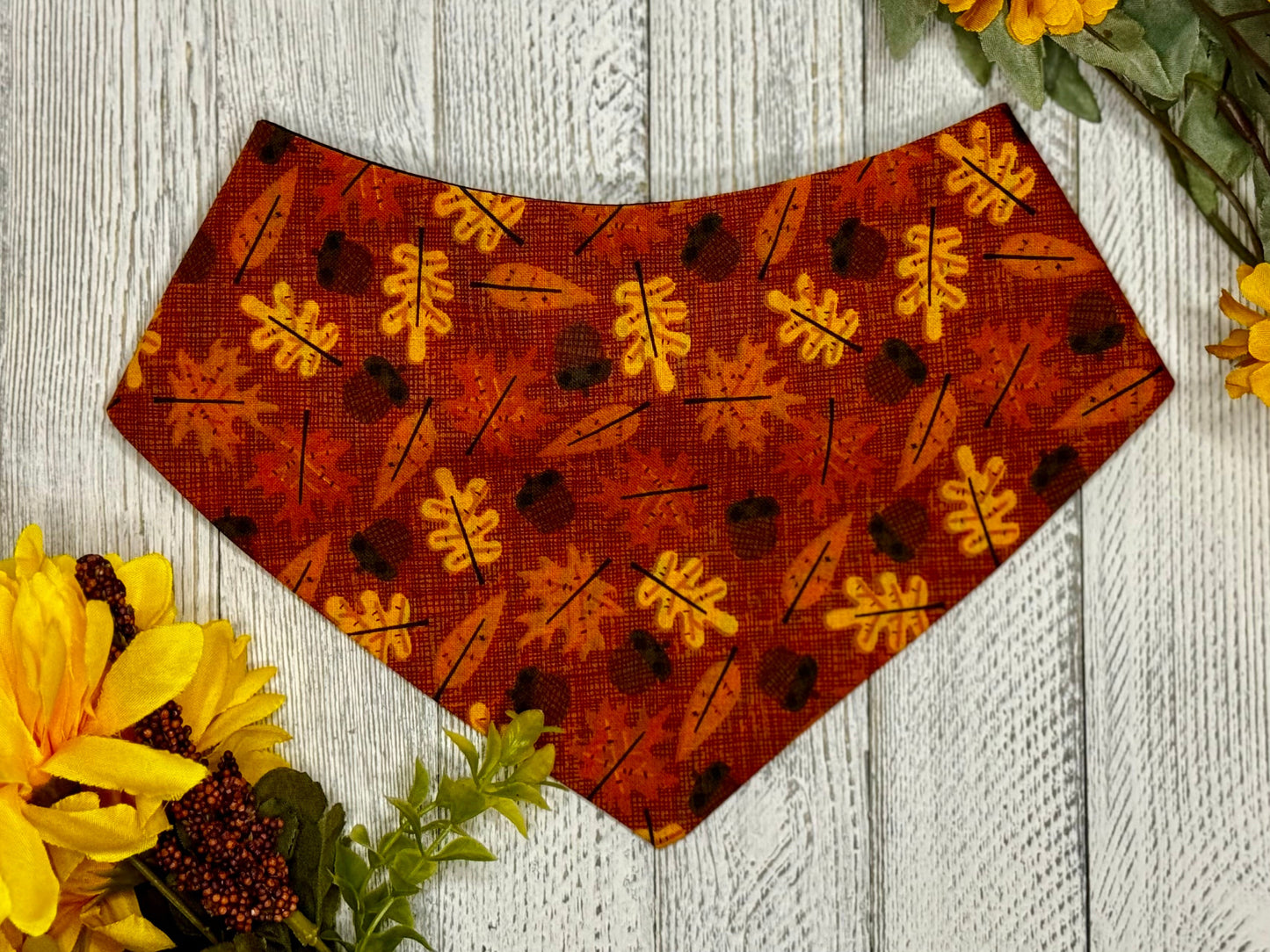 A scrunchie-style pet bandana with a leaf pattern in shades of brown and orange, displayed on a white wooden background with fall flowers around it.