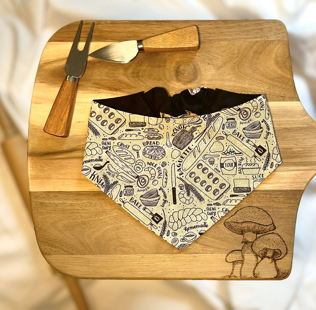This picture shows a scrunchie bandana sitting on a cutting board with cheese knifes on the side. There are mushrooms burned into the wood and the bandana displays baking related items like bread, rolling pin, muffin tin, and spatula. 