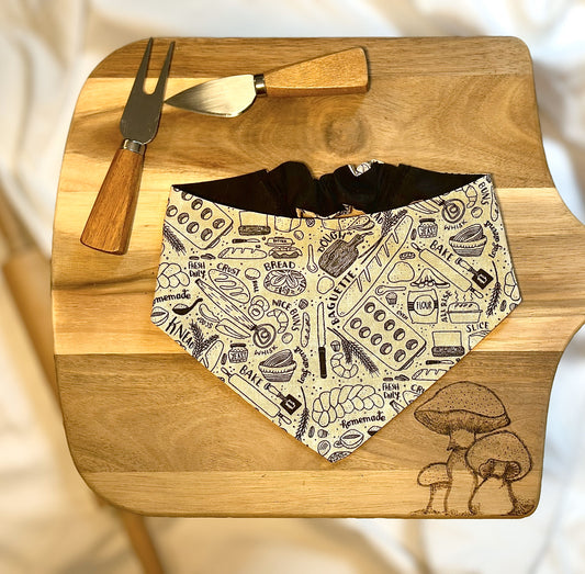 This picture shows a scrunchie bandana sitting on a cutting board with cheese knifes on the side. There are mushrooms burned into the wood and the bandana displays baking related items like bread, rolling pin, muffin tin, and spatula. 