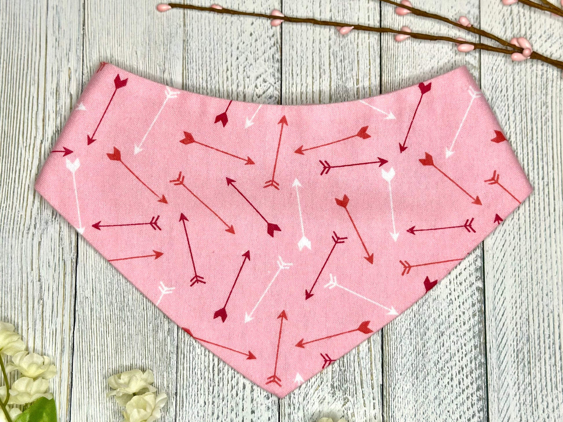 A pet bandana with a pink background featuring a red and white arrows, displayed on a wooden surface with flowers for decoration. 