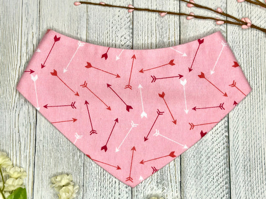 A pet bandana with a pink background featuring a red and white arrows, displayed on a wooden surface with flowers for decoration. 