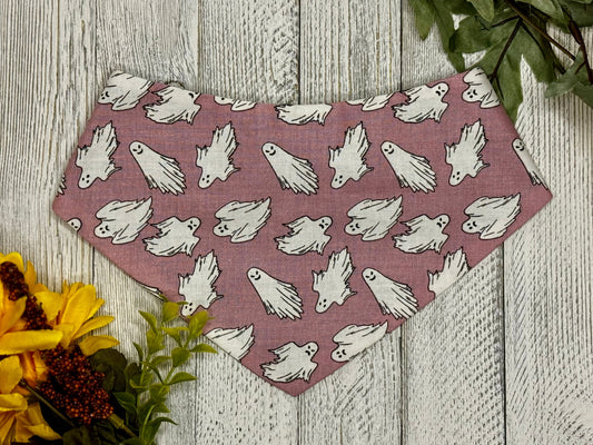 A pink scrunchie bandana with a pattern of white ghosts displayed on a wooden background with flowers and a green leaf.