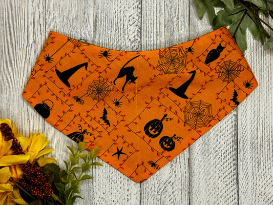 A pet bandana with a Halloween theme, featuring black cats and cobwebs on an orange background, displayed on a wooden surface with autumn leaves around it.