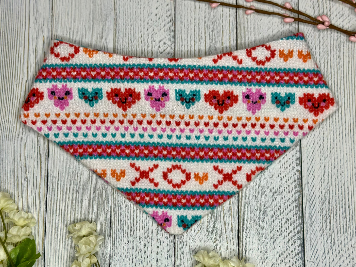 A pet bandana with a white background featuring a XOXO pattern and smiling hearts in red, pink, and blue, displayed on a wooden surface with flowers for decoration. 