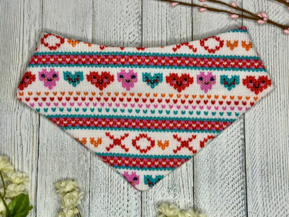 A pet bandana with a white background featuring a XOXO pattern and smiling hearts in red, pink, and blue, displayed on a wooden surface with flowers for decoration. 