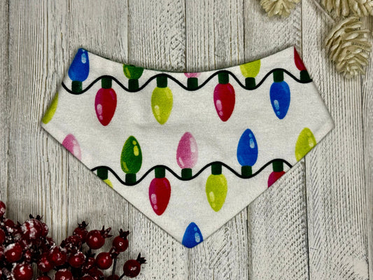 A pet bandana with a Christmas lights pattern displayed on a wooden background, surrounded by decorative elements.
