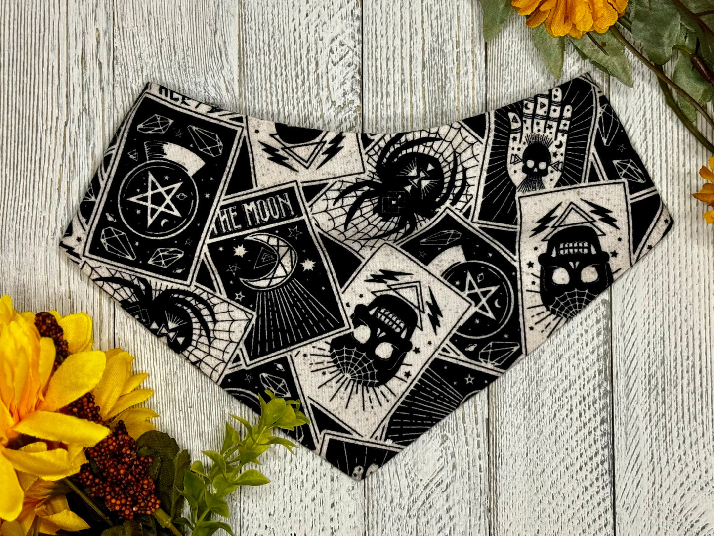 A black bandana with white designs, laid out on a wooden surface surrounded by yellow flowers. The bandana features a mystical and tarot card-like pattern.