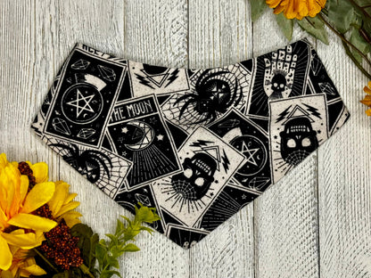 A black bandana with white designs, laid out on a wooden surface surrounded by yellow flowers. The bandana features a mystical and tarot card-like pattern.