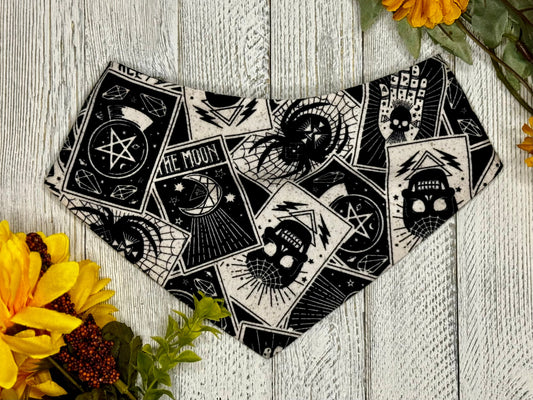 A black bandana with white designs, laid out on a wooden surface surrounded by yellow flowers. The bandana features a mystical and tarot card-like pattern.