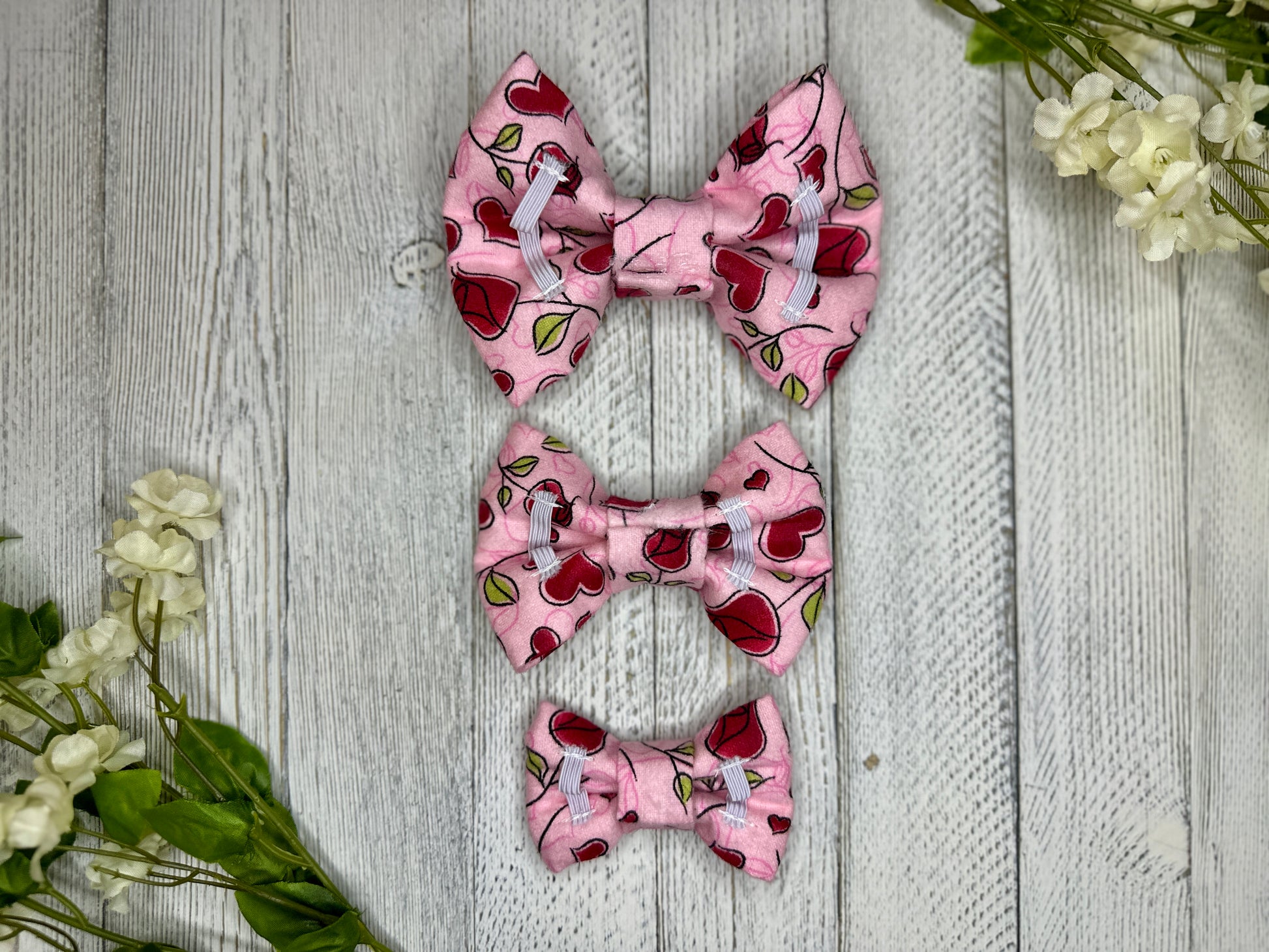 Picture of the backside of a pet bow tie showing the elastic straps that fit around a collar. Displayed on a wooden surface with flowers for decoration. 