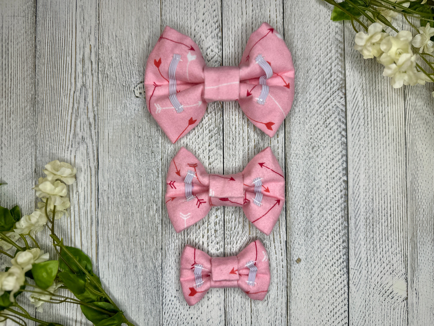 Picture of the backsides of three pet bow ties showing the elastic straps that fit around a collar. Displayed on a wooden surface with flowers for decoration. 