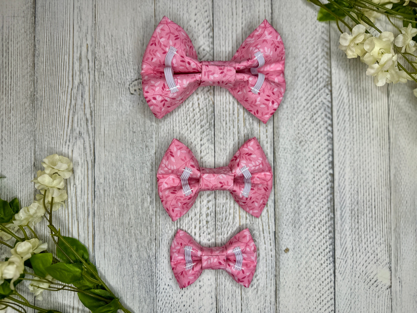 Picture of the backside of pet bow ties showing the elastic straps that fit around a collar. Displayed on a wooden surface with flowers for decoration. 