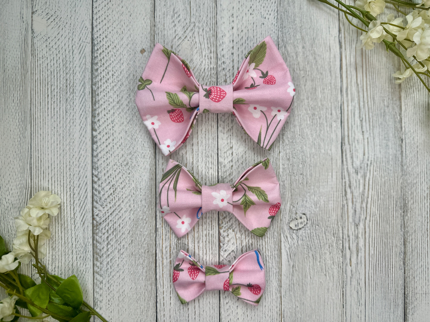 Three pet bow ties (small, medium, large) with a pink background and strawberries, displayed on a wooden surface with flowers for decoration. 