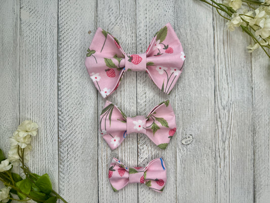 Three pet bow ties (small, medium, large) with a pink background and strawberries, displayed on a wooden surface with flowers for decoration. 