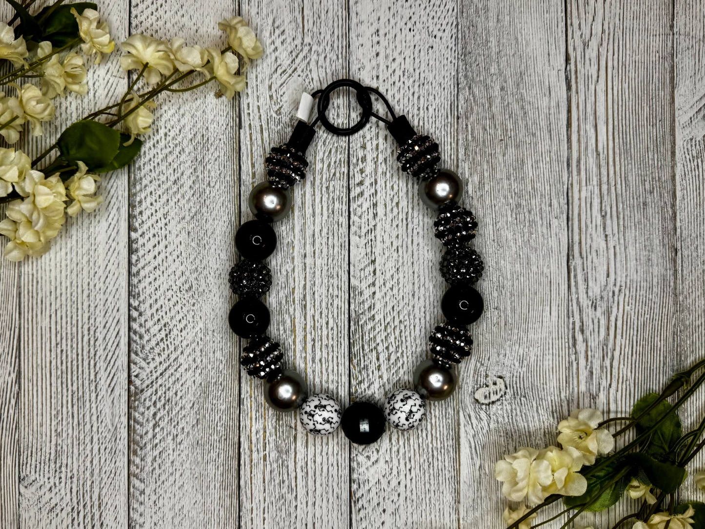 Beaded dog necklace with a black and white pattern including skeletons. Displayed on a wooden surface with flowers for decoration.