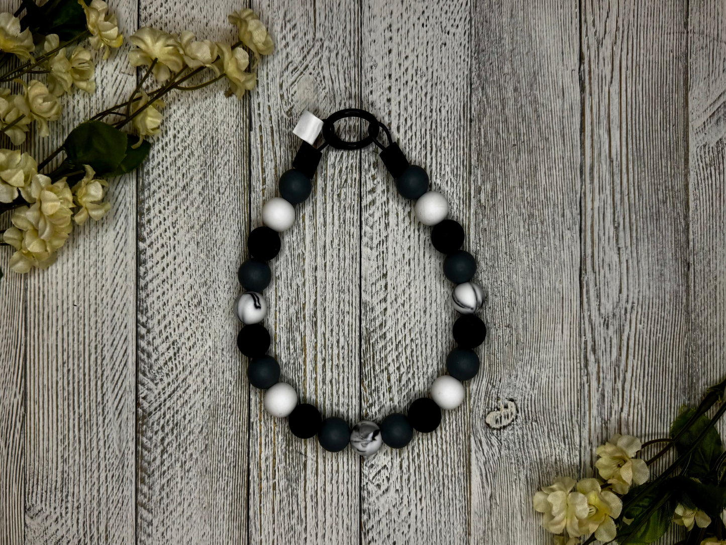 Beaded dog necklace with a black and white pattern including a marble effect. Displayed on a wooden surface with flowers for decoration.
