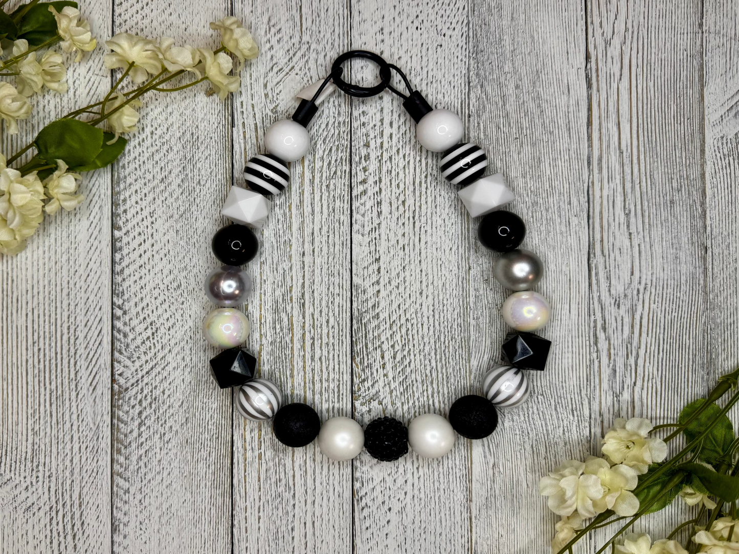 Beaded dog necklace with a black and white pattern including stripes. Displayed on a wooden surface with flowers for decoration.