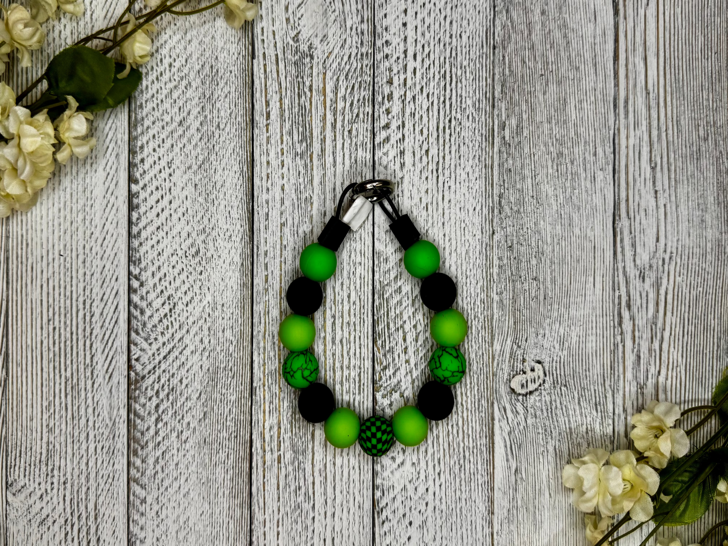 Beaded dog necklace with green and black pattern including checkered and cracked. Displayed on a wooden surface with flowers for decoration