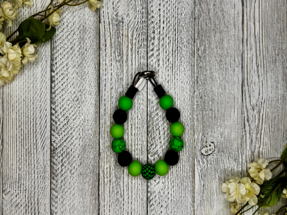 Beaded dog necklace with green and black pattern including checkered and cracked. Displayed on a wooden surface with flowers for decoration
