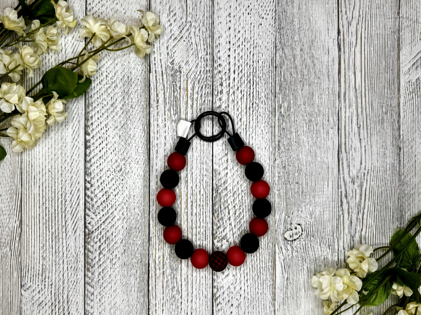 Beaded dog necklace with a red and black pattern including checkers. Displayed on a wooden surface with flowers for decoration.