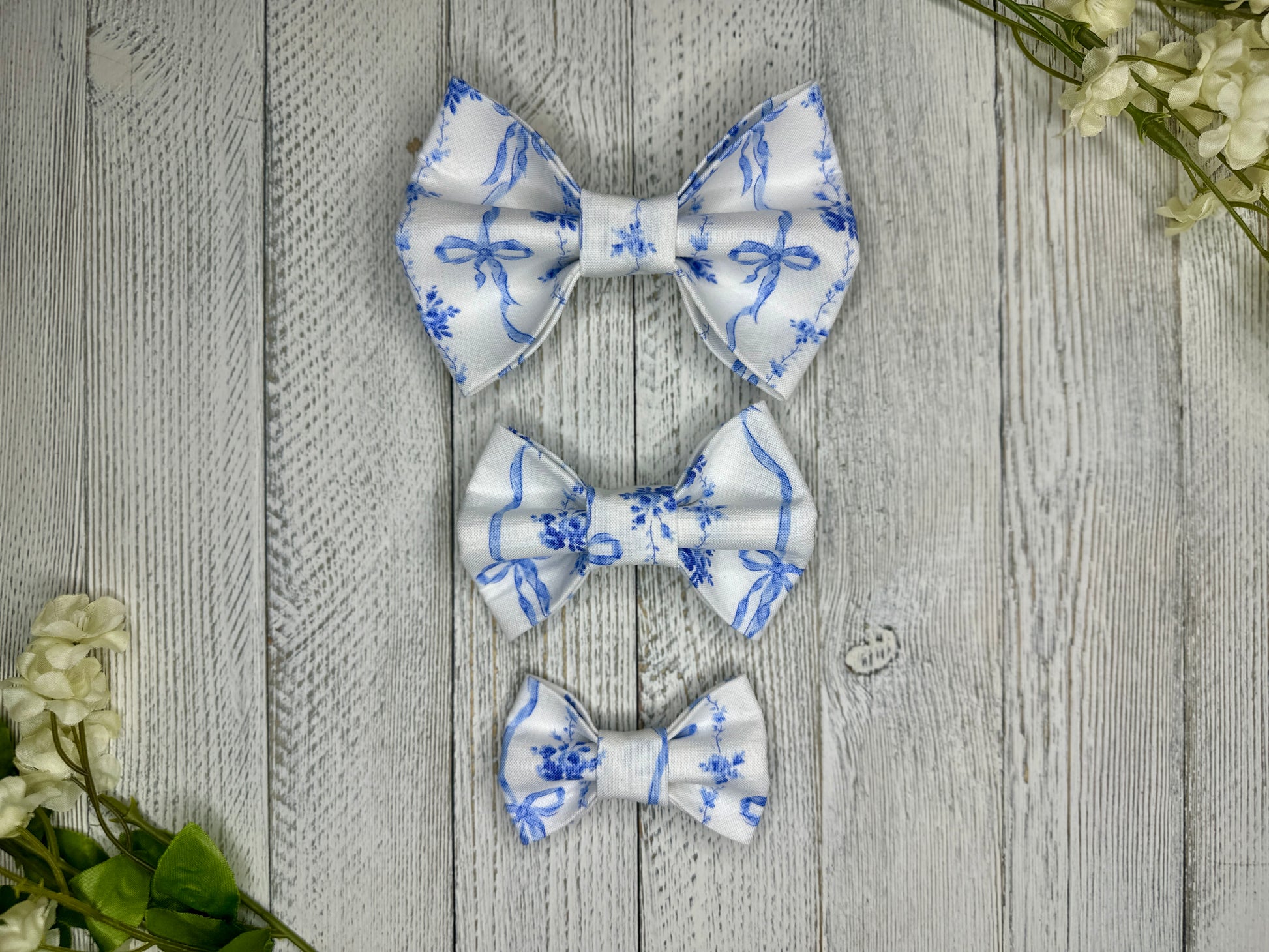 Three pet bow ties (small, medium, large) with a white background and blue bow pattern displayed on a wooden surface with flowers for decoration. 