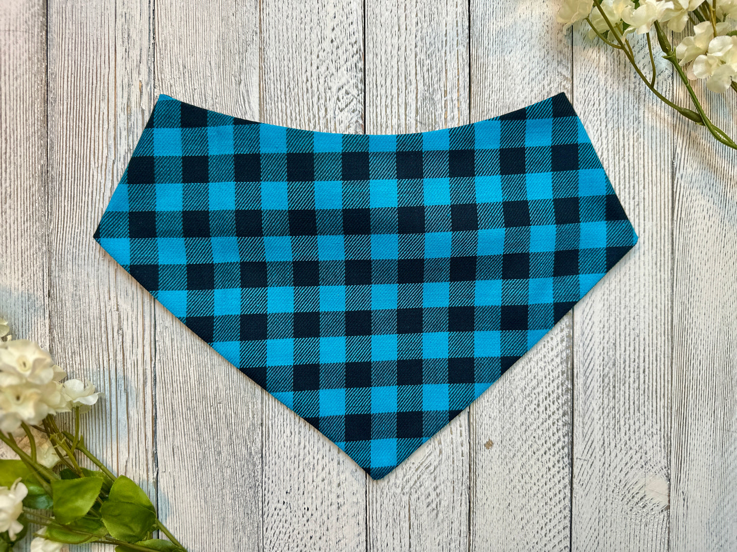 A pet bandana with a blue and black checkered pattern displayed on a wooden surface with flowers for decoration. 