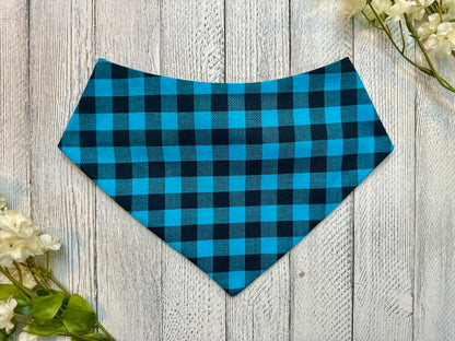 A pet bandana with a blue and black checkered pattern displayed on a wooden surface with flowers for decoration. 