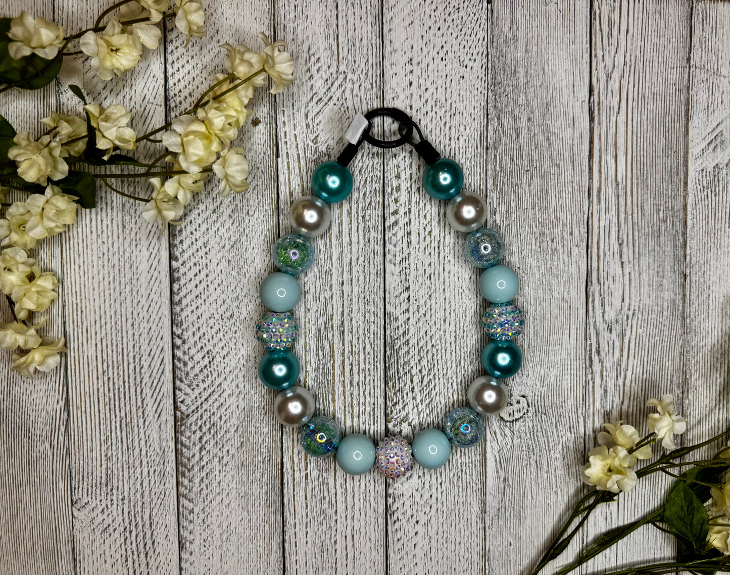 Beaded dog necklace with a light blue and silver pattern including stripes. Displayed on a wooden surface with flowers for decoration.