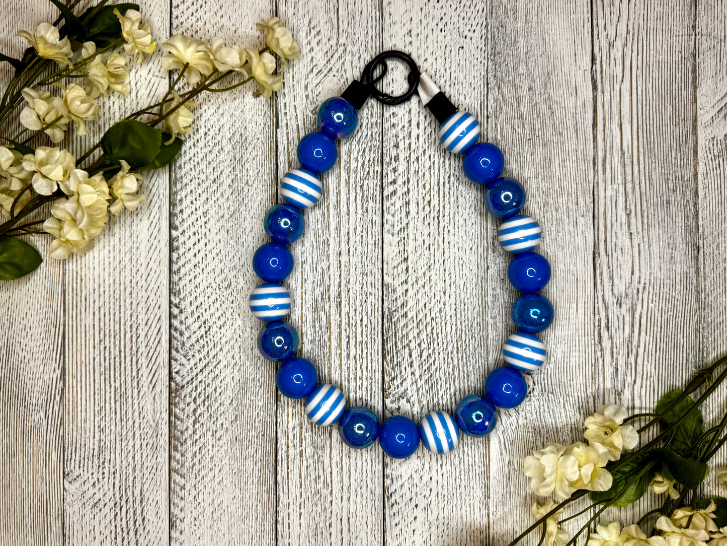 Beaded dog necklace with a blue and white pattern including stripes. Displayed on a wooden surface with flowers for decoration.