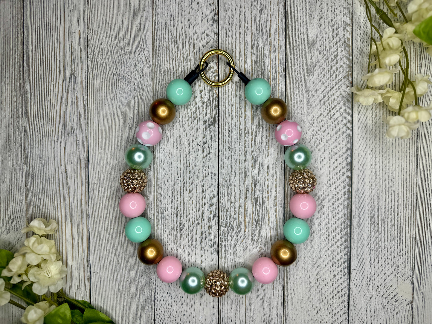 Beaded pet necklace with a gold, pink, and teal pattern, including polka dots. Displayed on a wooden surface with flowers for decoration. 