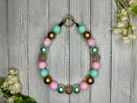 Beaded pet necklace with a gold, pink, and teal pattern, including polka dots. Displayed on a wooden surface with flowers for decoration. 