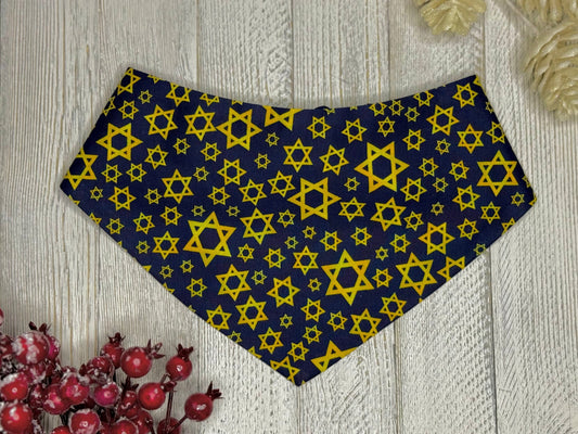 A pet bandana featuring a blue background with yellow Star of David patterns, displayed on a wooden surface with decorative berries.