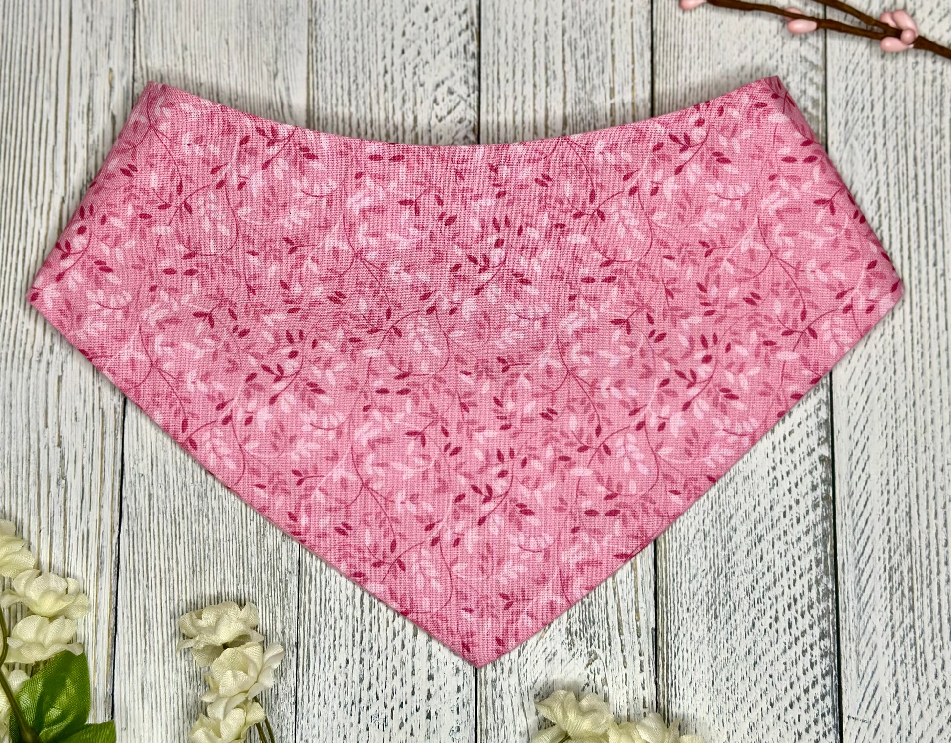 A pet bandana with a pink background featuring a subtle leaf design in light and dark pink, displayed on a wooden surface with flowers for decoration. 