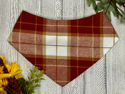 A checkered bandana in orange, red, and beige colors, designed for pets, displayed on a wooden background with fall-themed decorations.
