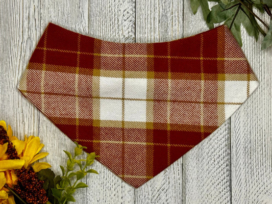 A checkered bandana in orange, red, and beige colors, designed for pets, displayed on a wooden background with fall-themed decorations.