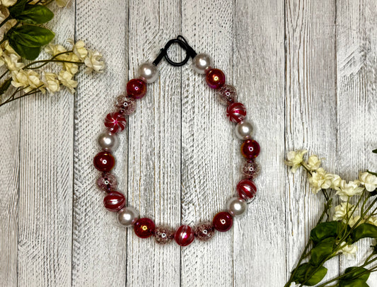 Beaded dog necklace with a red and white pattern including candy cane swirls. Displayed on a wooden surface with flowers for decoration.