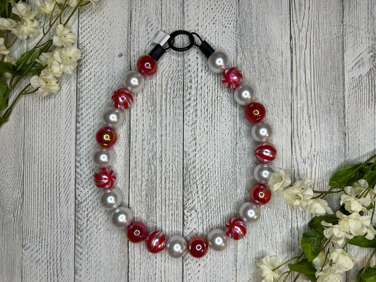 Beaded dog necklace with a red and white pattern including candy cane swirls. Displayed on a wooden surface with flowers for decoration.