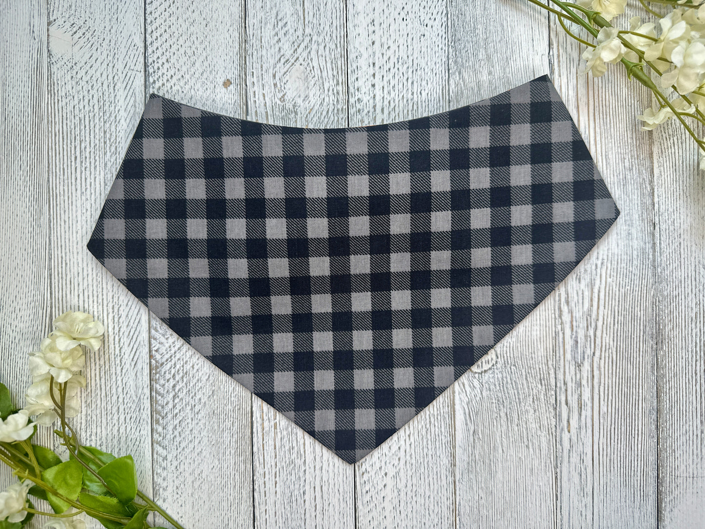 A pet bandana with a black and gray checkered pattern, displayed on a wooden surface with flowers for decoration. 