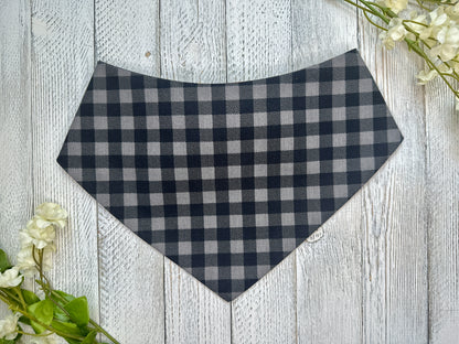 A pet bandana with a black and gray checkered pattern, displayed on a wooden surface with flowers for decoration. 