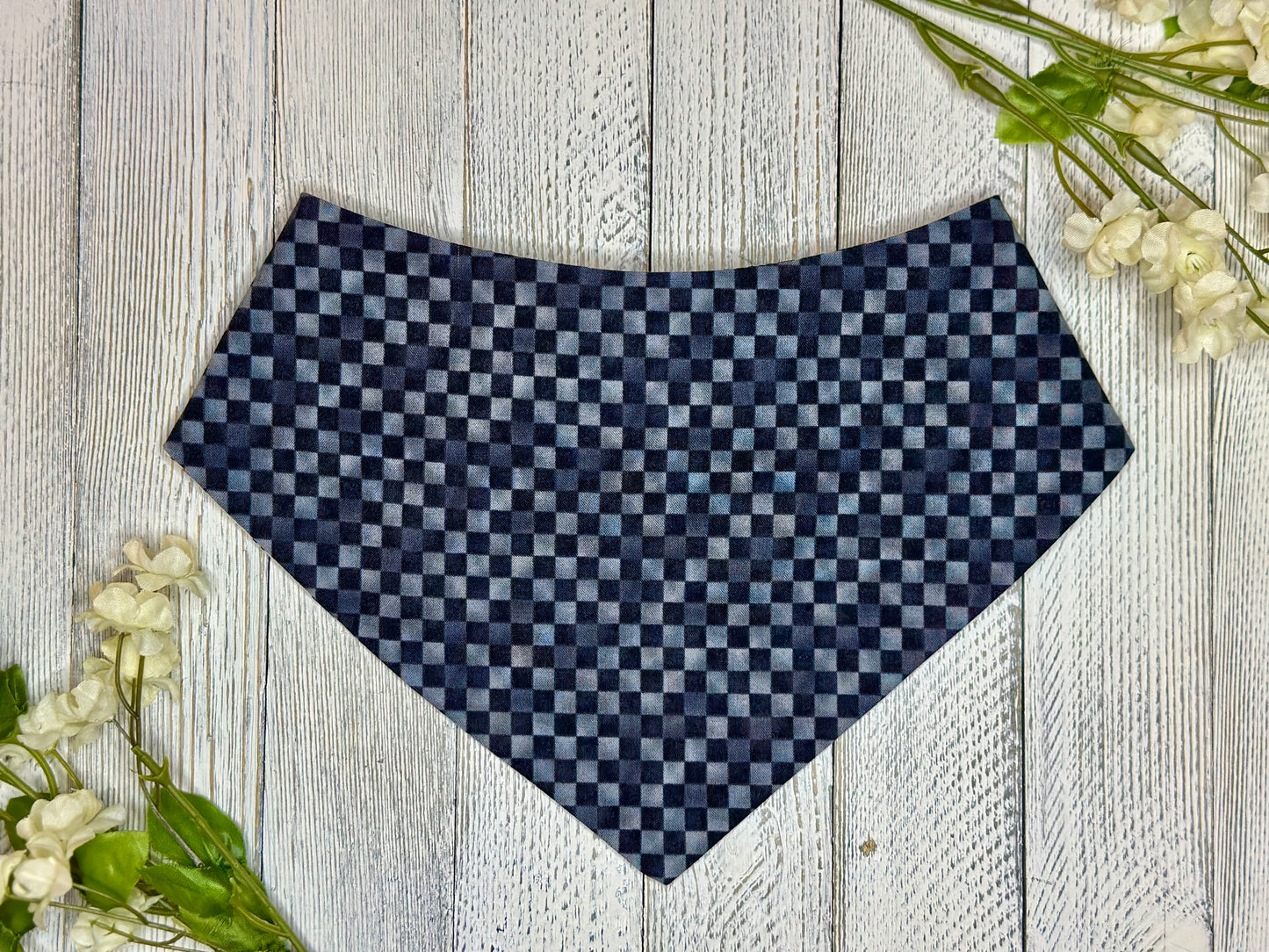 A pet bandana black and grey checkered pattern, displayed on a wooden surface with flowers for decoration. 