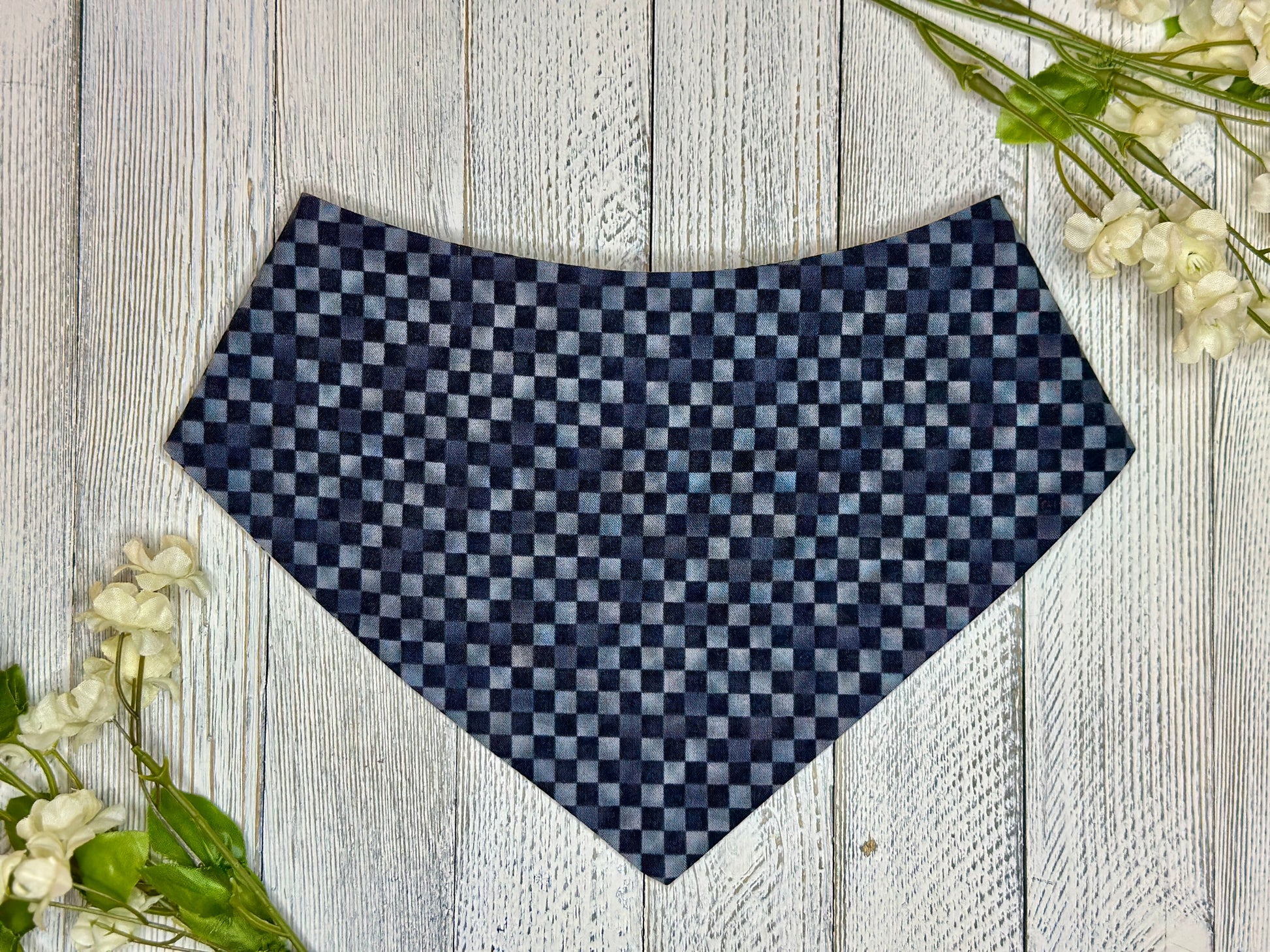 A pet bandana black and grey checkered pattern, displayed on a wooden surface with flowers for decoration. 