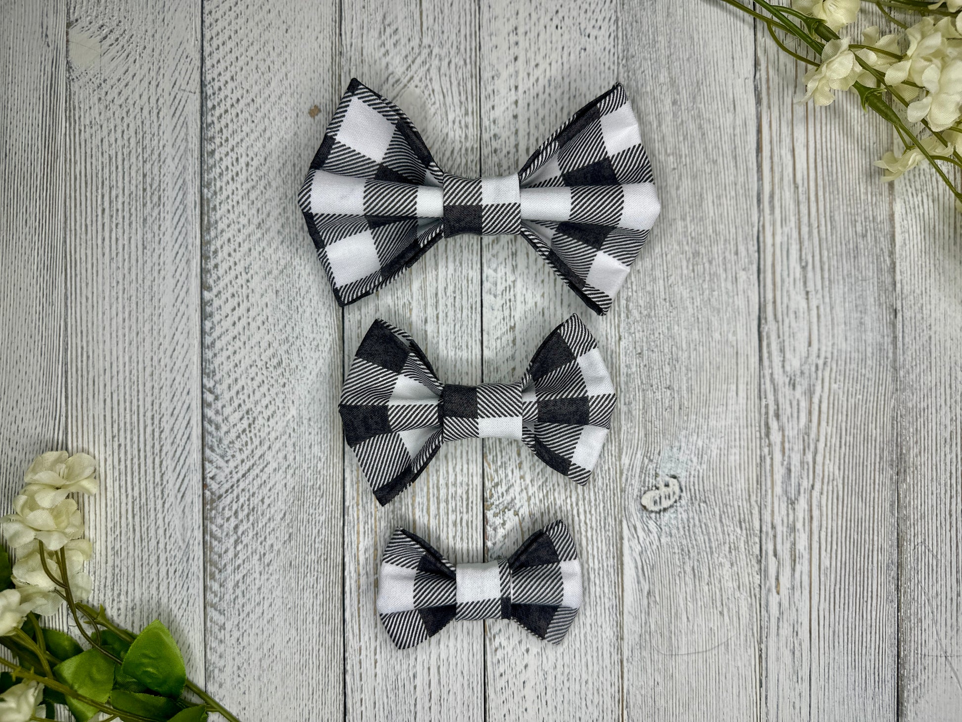 Three pet bow ties (small, medium, large) with gray and white checkered pattern displayed on a wooden surface with flowers for decoration. 