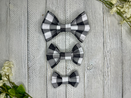 Three pet bow ties (small, medium, large) with gray and white checkered pattern displayed on a wooden surface with flowers for decoration. 