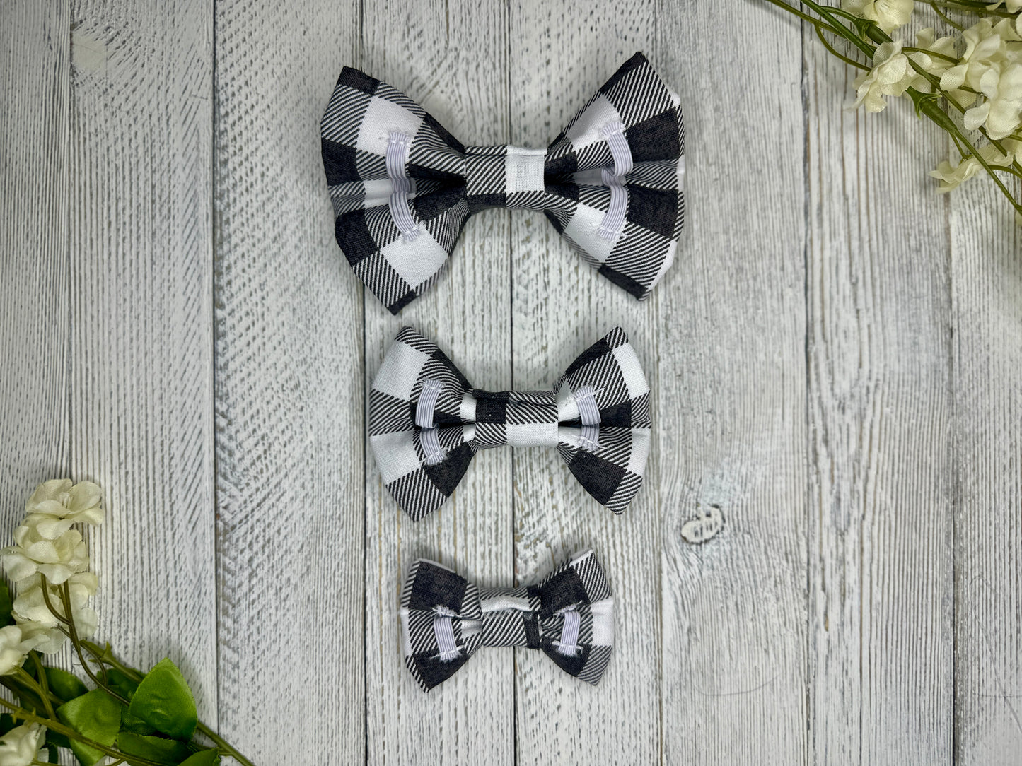 Picture of the backside of pet bow ties showing the elastic straps that fit around a collar. Displayed on a wooden surface with flowers for decoration. 