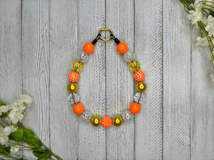 Beaded pet necklace with a yellow and orange pattern, including polka dots. Displayed on a wooden surface with flowers for decoration. 