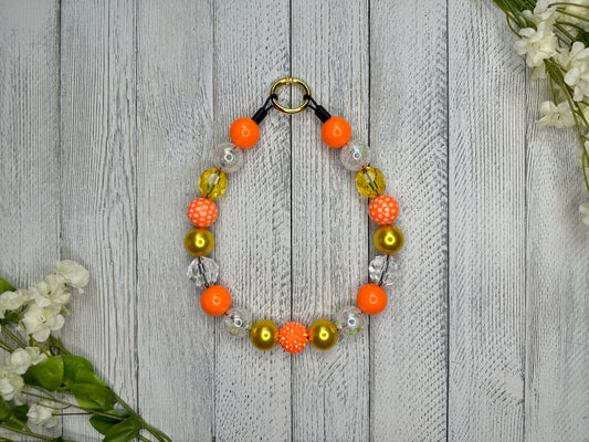 Beaded pet necklace with a yellow and orange pattern, including polka dots. Displayed on a wooden surface with flowers for decoration. 