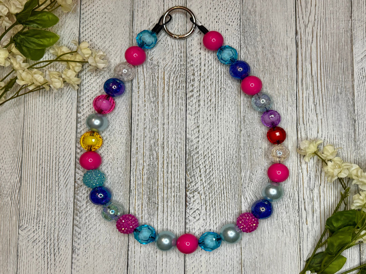 Beaded pet necklace with a purple, blue, and pink pattern. Displayed on a wooden surface with flowers for decoration. 
