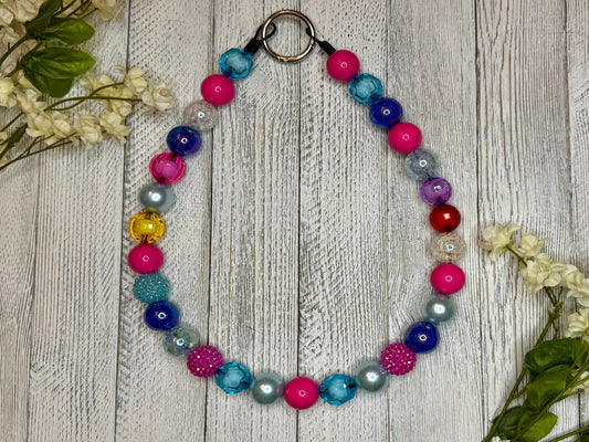 Beaded pet necklace with a purple, blue, and pink pattern. Displayed on a wooden surface with flowers for decoration. 