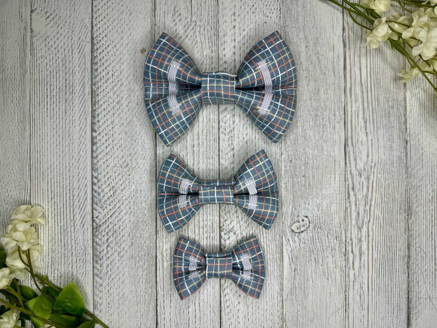 Picture of the backside of pet bow ties showing the elastic straps that fit around a collar. Displayed on a wooden surface with flowers for decoration. 