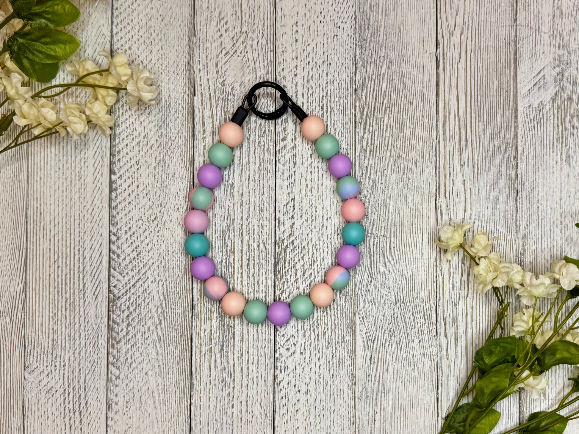 Beaded dog necklace with a pink, blue, and purple pattern. Displayed on a wooden surface with flowers for decoration.